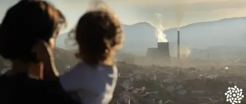 Mãe com filha no colo observa fumaça saindo de chaminé industrial à distância. A poluição é um dos fatores de atenção quando o tema é a exposição ambiental de crianças